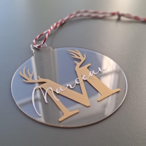 Peut inclure: Ornement de Noël en acrylique transparent avec la lettre "M" dorée et des bois de cerf. Le nom "Marceau" est écrit en blanc. Une boucle en ficelle rouge et blanche est attachée pour l'accrocher.