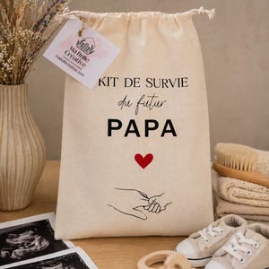 Peut inclure: Un pochon en coton beige avec cordon de serrage portant l'inscription noire "KIT DE SURVIE du futur PAPA", un cœur rouge et un dessin au trait représentant une main d'adulte tenant une main de bébé. Une étiquette "Ma Bulle Créative" est attachée.