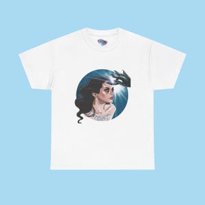 May include: White t-shirt featuring a circular graphic of a woman with dark hair, a hand reaching towards her head, and the text "MAGICAL LUST". The graphic is set against a blue background.