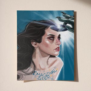 May include: Art print featuring a woman with dark hair and red lipstick, with the text "IMAGINAL DISK". A green claw-like hand hovers above her head, holding a reflective disc, with a bright light emanating from it. The artwork has a surreal, dreamlike quality.