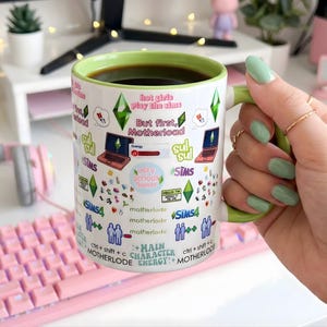 May include: A white ceramic mug with a green handle and rim, filled with black coffee. The mug is decorated with various graphics and text related to the Sims video game, including laptops, diamonds, and the words "hot girls play the sims."