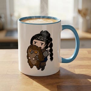 May include: White ceramic mug with a blue handle and rim, featuring a cartoon illustration of a character holding a brown owl. The mug contains a latte with a decorative design on top. The mug is on a wooden surface.