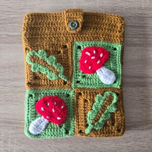May include: A brown crocheted pouch with a green button closure. The pouch is decorated with four squares, each featuring a different design: a green leaf, a red and white mushroom, a green leaf, and a red and white mushroom.