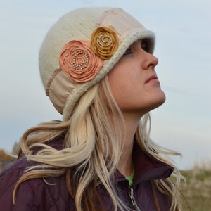 May include: A woman with blonde hair is wearing a white knitted hat with a brown and pink flower embellishment. She is wearing a maroon quilted jacket.