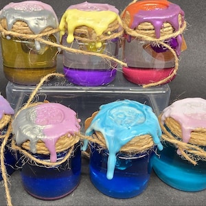 Op de afbeelding: Een verzameling kleine glazen potjes met gekleurde vloeistof. Elk potje is afgedekt met een decoratief deksel en touw. De deksels zijn beschilderd met druipende metallic en pastelkleuren, wat een grillige esthetiek creëert.