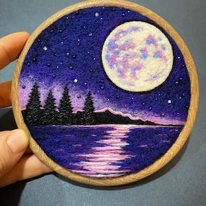 Puede incluir: Arte de bordado cosido a mano en un aro de madera. La obra de arte representa un paisaje nocturno con una luna llena, una masa de agua reflectante y árboles silueteados. La paleta de colores incluye tonos de morado, azul y rosa.