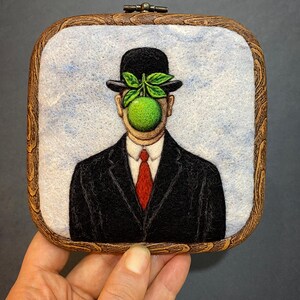 May include: A felt art piece depicting a man in a black suit, bowler hat, and red tie, with a green apple obscuring his face. The artwork is framed in a square wooden hoop with a decorative edge, and a light blue background.