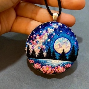 May include: Round pendant with a night sky design. The pendant features a blue background with a full moon, trees, and pink and white accents. It hangs from a black cord with a silver-colored clasp.