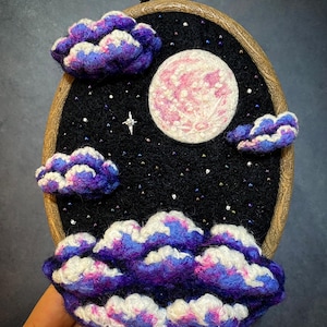 Op de afbeelding: Een ovale borduurring met een nachtscène. Het kunstwerk bevat een roze en witte maan, pluizige paarse en witte wolken en verspreide sterren. De achtergrond is zwart en de ring is van hout.