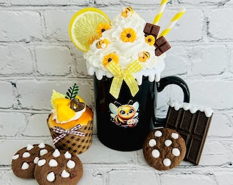 May include: A black mug with a bee graphic, topped with whipped cream, lemon slice, and decorative bees and flowers. Includes a cupcake, chocolate chip cookies, and a chocolate bar. Yellow straws and bow add to the whimsical design.
