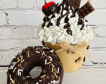 May include: A decorative dessert display featuring a chocolate-covered donut with sprinkles and a sundae. The sundae has whipped cream, chocolate syrup, chocolate pieces, a cherry, and wafer sticks. The sundae is in a small paper cup.