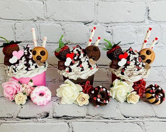 May include: Three decorative faux milkshakes with whipped cream, chocolate, and strawberry toppings. Each milkshake has a straw, a cookie, and a strawberry. Pink, brown, and red cups are decorated with flowers and donuts.