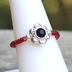 May include: A ring featuring a central silver-toned flower-shaped charm with a small black circular stone at its center. The band of the ring is constructed from a series of small, translucent red beads strung together on a flexible wire.