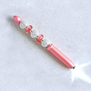 May include: A pink pen featuring a series of decorative beads along the upper barrel, including two textured silver-toned spheres and two translucent pink patterned beads, with a bright white LED light emitting from the tip of the pen.