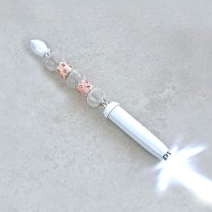 May include: A white pen with a light-up tip emitting a bright white glow. The handle features a decorative sequence of alternating silver-toned mesh spheres and rose gold-toned cylindrical beads, topped with a smooth white pointed finial.