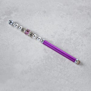 May include: A purple stylus featuring a decorative handle adorned with a series of silver-toned beads and a central cylindrical bead with pink accents. The tip of the stylus is rounded and light-colored, designed for use on touchscreens.