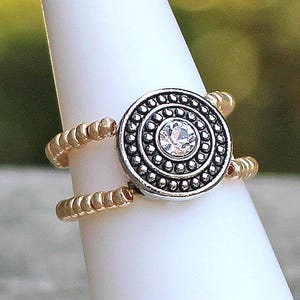 May include: A silver and gold ring with a central circular design. The centre of the ring features a clear, round gemstone surrounded by small silver beads. The band is made of gold-coloured beads, creating a two-part design.
