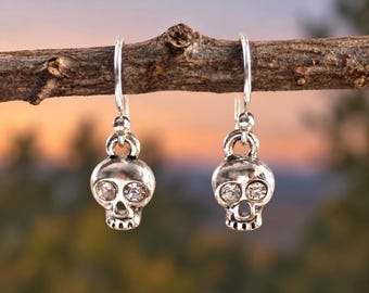 May include: A pair of silver-toned drop earrings featuring a skull design. Each skull charm is crafted with a polished metallic finish and includes two clear, round faceted crystals set into the eye sockets, suspended from simple silver-toned hooks.