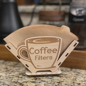 以下が含まれることがあります： コーヒーカップと「Coffee Filters」の文字がレーザーカットされた木製のコーヒーフィルターホルダー。ホルダーには茶色の紙フィルターが入れられ、カウンタートップに置かれています。