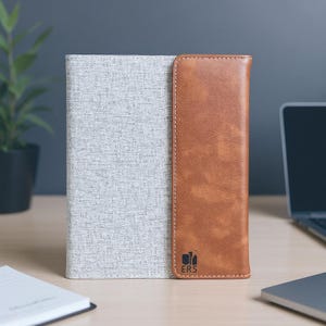 May include: A gray fabric and brown leather book cover. The cover has a light gray textured fabric on the left side and a brown leather flap on the right side. The brown leather flap has the letters "ERS" printed on it.