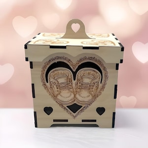 May include: A light-colored wooden box with a heart-shaped handle and cutouts. The front features a heart-shaped design with two baby shoes. The box has a square shape and is likely for storage or decoration.