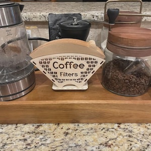 Könnte beinhalten: Ein hellbrauner Holz-Kaffeefilterhalter mit den Worten "Coffee Filters" ausgeschnitten. Der Halter steht auf einem weißen Sockel. Ein Glaskessel, eine Kaffeemühle und Gläser mit Kaffeebohnen sind ebenfalls zu sehen.