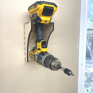 May include: A yellow and black cordless drill mounted on a wooden wall mount. The drill is held in place by a slot in the mount. The drill has a black and silver handle and a black bit.