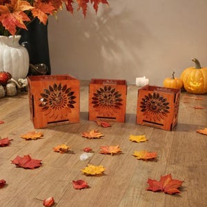 Könnte beinhalten: Drei hölzerne Kerzenhalter mit Sonnenblumen-Ausschnitten, angeordnet auf einem Holzboden, der mit Herbstblättern übersät ist. Eine Vase mit Herbstlaub und geschnitzten Kürbissen befindet sich im Hintergrund und schafft eine saisonale Atmosphäre.