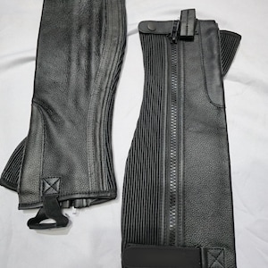 May include: Two black leather riding chaps. One chap is open, revealing a zipper closure, while the other is closed. Both chaps have a textured surface and vertical ribbed detailing. Each chap has a buckle and strap closure.
