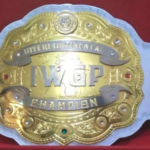 Könnte beinhalten: Ein Meisterschaftsgürtel mit einer großen, vergoldeten Mittelplatte, die den Schriftzug IWGP INTERCONTINENTAL CHAMPION in silbernen Buchstaben trägt. Der Gürtel verfügt über weiße Lederriemen, dekorative Seitenplatten sowie kleine rote und grüne Edelsteine.