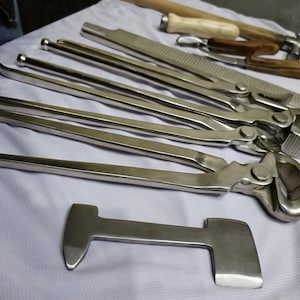 May include: A set of nine silver metal farrier tools, including a rasp, a hammer, and a variety of nippers. The tools are laid out on a white surface.