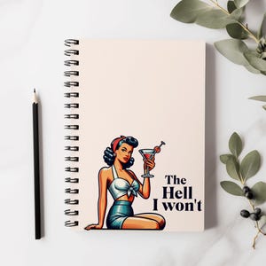 Op de afbeelding: Een spiraalgebonden notitieboek met een witte omslag met een cartoonillustratie van een vrouw die een cocktailglas vasthoudt. De tekst "The Hell I won't" is in zwart onder de illustratie gedrukt.