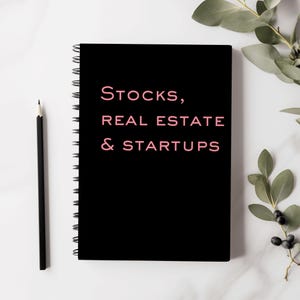 Op de afbeelding: Een zwarte spiraalgebonden notitieboek met de tekst "Stocks, Real Estate & Startups" in roze letters.