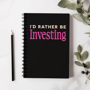 Può includere: Quaderno nero a spirale con la scritta "I'D RATHER BE Investing" in lettere bianche e rosa. Una matita nera è a sinistra del quaderno. C'è del verde nell'angolo in alto a destra. Il quaderno è su una superficie di marmo bianco.
