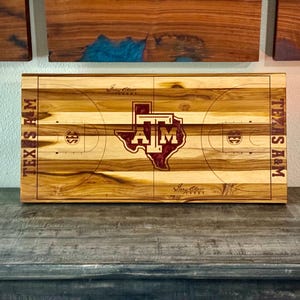 Puede incluir: Una tabla de cortar de madera con un diseño de cancha de baloncesto y el logotipo de Texas A&M en granate. La tabla está etiquetada "Texas A&M" en ambos lados.