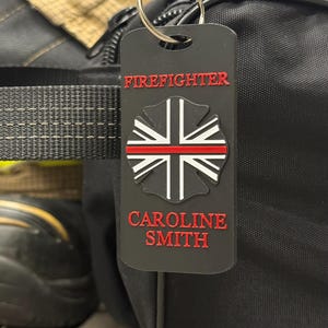 Puede incluir: Etiqueta rectangular negra con las palabras "FIREFIGHTER" y "CAROLINE SMITH" en rojo. La etiqueta presenta un emblema de bombero con una línea roja. Está sujeta a una bolsa negra con un llavero plateado.