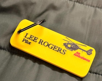 Puede incluir: Una etiqueta rectangular de color amarillo brillante con el nombre "LEE ROGERS" y la palabra "Pilot" impresos en negro. Un gráfico de helicóptero negro y rojo con las palabras "Air Ambulance" también está impreso en la etiqueta. Un clip de metal negro está adjunto.