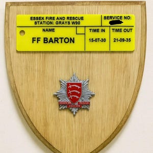 May include: A wooden plaque with a yellow name tag and a silver emblem. The name tag reads "ESSEX FIRE AND RESCUE" and "FF BARTON". The emblem features a red shield with three curved lines and the words "ESSEN ER RESUE ENCLARE".