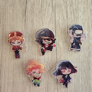 May include: Five acrylic charms featuring anime-style characters. The characters have various hairstyles and outfits, with a range of colors including red, black, blue, and gold. The charms are displayed on a light wood-grain surface.