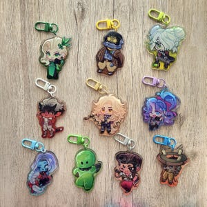 May include: A collection of colorful acrylic keychains featuring various anime-style characters. Each keychain has a unique design with vibrant colors and attached metal clasps for easy attachment to bags or keys. The keychains are displayed on a wooden surface.