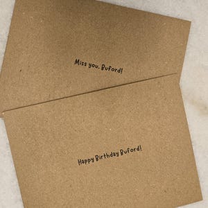May include: Two brown greeting cards with black text. The top card reads "Miss you, Buford!" and the bottom card reads "Happy Birthday Buford!" The cards are made of a textured, recycled paper.