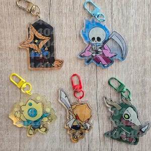 May include: Five acrylic keychains featuring cartoon characters: a star-shaped creature in a dark frame, a skeleton with a scythe and blue flames, a yellow creature with a blue cape, a gold-armored warrior with a sword, and a green hooded figure.