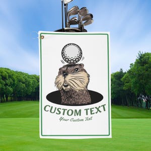 May include: White golf towel with a green border and a cartoon groundhog design. The groundhog is looking up at a golf ball. The towel has the words "CUSTOM TEXT" and "Your Custom Text" in green.