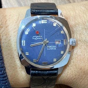 May include: A vintage Mortima Super 26 wristwatch with a square stainless steel case and a dark blue dial. The watch features a black leather band and a date display at the 3 o'clock position. The dial has silver-tone hour markers and hands.