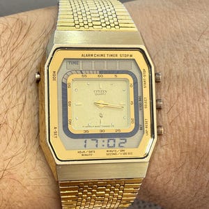 Op de afbeelding: Een goudkleurig Citizen digitaal horloge met een rechthoekige wijzerplaat en bijpassende band. De tijd wordt weergegeven als 17:02. De wijzerplaat bevat de tekst "CITIZEN QUARTZ" en functielabels. Het horloge zit om een pols.