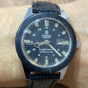 May include: A vintage Mortima Super 28 automatic wristwatch with a black bezel and a black leather band. The watch face is black with white and green markers, and the date is displayed at the 3 o'clock position. The watch is waterproof.