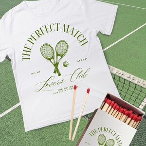 Könnte beinhalten: Weißes T-Shirt mit Tennis-Design, einschließlich gekreuzter Schläger, Ball und dem Text "The Perfect Match Lovers Club". Eine Streichholzschachtel und Streichhölzer sind ebenfalls zu sehen.