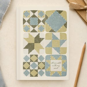 Puede incluir: Un cuaderno rectangular con cubierta blanca que presenta seis bloques cuadrados estilo colcha en tonos verde salvia, oliva y azul polvoriento. Los bloques muestran patrones geométricos con detalles de costura blancos. El bloque inferior derecho contiene el texto It is well with my soul.