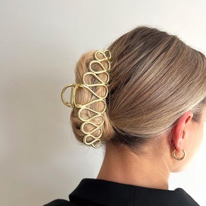 May include: A gold-colored hair claw clip with a unique looped design, holding up a bun. The clip is made of metal and has a shiny finish. The word "Dreamy" is in the top left corner.