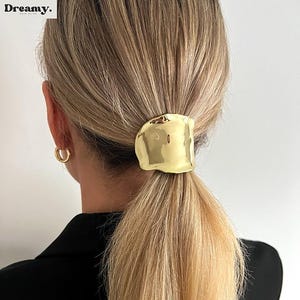Puede incluir: Una goma para el pelo de metal dorado con una forma ondulada y orgánica. La goma se lleva en una coleta.
